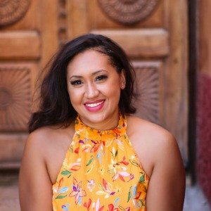 Taylor Shuler, smiling, wearing an orange halter top with brightly colored flowers in front of a carved wooden front door.